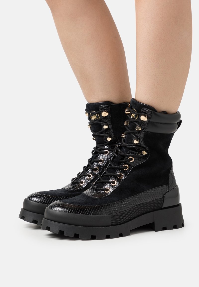 Bottes de combat noires à lacets avec semelles épaisses et garnitures dorées portées sur des jambes nues.