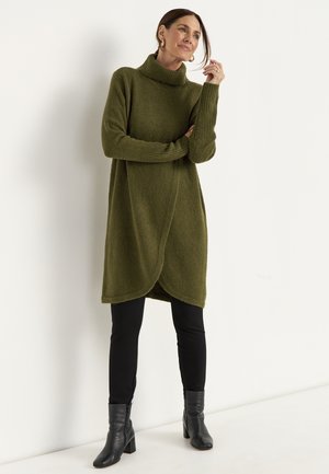 Kvinde iført en olivengrøn rulneck-sweaterkjole, sorte leggings og sorte ankelstøvler, står mod en ensfarvet lys væg.