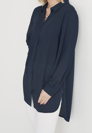 Femme portant une longue chemise bleu marine boutonnée avec un col et un pantalon blanc, debout contre un mur blanc.