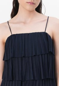Robe plissée marine avec plusieurs volants en cascade, fines bretelles et texture douce. Design simple et fluide mettant l'accent sur les lignes verticales.