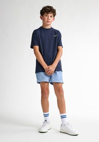 Camiseta de manga corta azul marino con un logo verde, combinada con pantalones cortos azul claro. El modelo lleva zapatillas blancas y calcetines a rayas.
