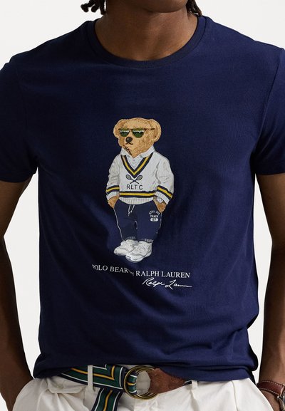 Marineblauwe katoenen t-shirt met een berenafbeelding in sportoutfit met zonnebril, geborduurd RLTC-logo en handtekening onder de illustratie.