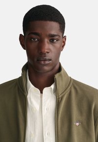 Chaqueta de cremallera verde oliva con cuello alto sobre una camisa blanca de botón. Con un pequeño logo colorido en el pecho. Textura suave.