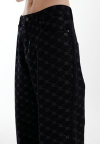 Pantalon en denim noir avec un motif ton sur ton texturé. Le design comprend des poches et une fermeture à l'avant avec un bouton métallique.
