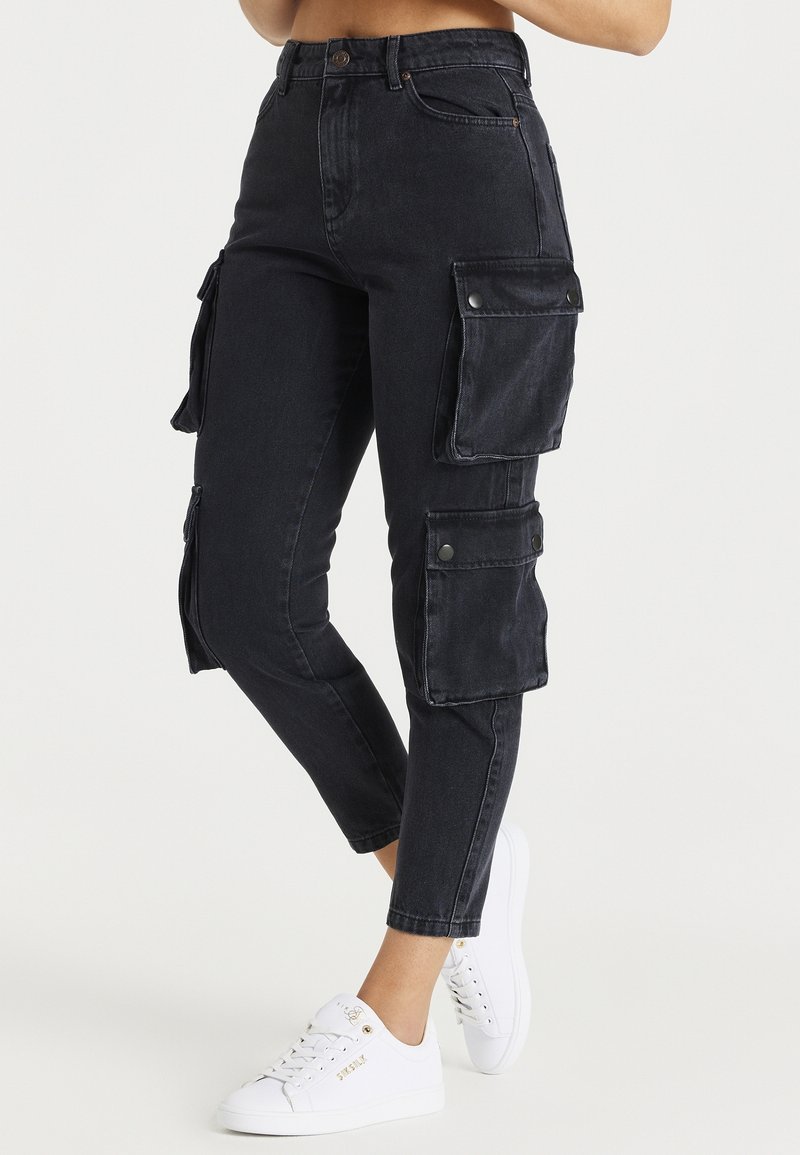 Personne portant un pantalon cargo noir avec plusieurs poches à rabat et des baskets blanches, debout devant un fond clair et uni.