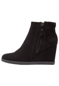 Bottines en suède noir à talon compensé et à fermeture éclair sur le côté, présentant une texture lisse, un bout rond et un talon discret pour une hauteur supplémentaire.