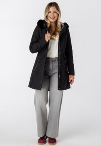 Zwarte wolmix jas met een nepbont capuchon, voorzijde met rits en zijzakken; gecombineerd met grijze wijdvallende jeans en bordeauxrode sneakers.