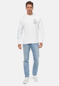 Hvid langærmet T-shirt med et tegneserieprint på brystet og farverige designs på ærmerne, parret med lyseblå jeans.