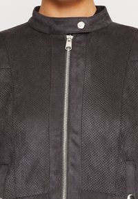 Giacca in finta suede perforata nera con colletto alto, chiusura con zip argentata e dettagli con bottoni a scatto. Superficie testurizzata con cuciture dettagliate.