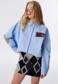 Camicetta azzurro chiaro corta con tasca a quadri e toppe sui gomiti; abbinata a una gonna a rombi neri e bianchi con dettagli argentati.