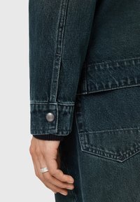 Tummanvihreä denimitakki, jossa on napilliset hihansuut, tekstuurikangas ja sivutasku. Kankaassa on hieman kuluneen näköinen pinta.