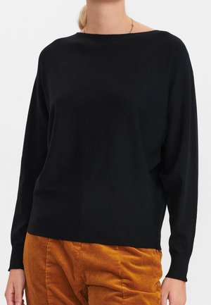 Femme portant un pull noir à manches longues et un pantalon en velours côtelé orange-brun, montrée du cou aux hanches sur un fond uni.