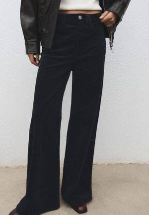 Pantalones de pana negros de pierna ancha, con una cintura alta, un solo botón en la parte delantera y relieves texturizados. Complementados con zapatos de un burdeos oscuro.