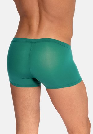 Close-up van het onderlichaam en de benen van een persoon die strakke, groene zwembroek draagt tegen een effen, lichte achtergrond.