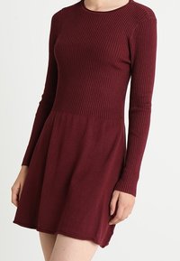 Abito bordeaux a maniche lunghe con texture a coste sulla parte superiore e una gonna a ruota. Il materiale sembra morbido e leggero. Collo rotondo semplice.