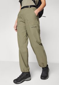 Olive zelene hlače za prosti čas imajo elastične manšete, stranske žepke in logo Columbia. Tkanina se zdi trdna in lahka. Vključeni so črni pohodniški čevlji.