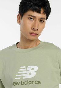 Sweatshirt vert clair à col rond en tissu doux, présentant un grand graphisme blanc du logo "New Balance" sur le devant.