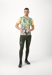 Zaļas sporta bikses ar teksturētu dizainu un pielāgotu dzeltenu un zilu grafisko T-kreklu, turēdams melnu un baltu futbola bumbu.