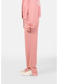 Sudadera rosa con cremallera, detalle de gran logo y pantalones de chándal rosa a juego. El calzado presenta un diseño texturizado en color blanco roto.