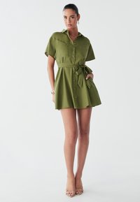 Abito chemisier verde corto con vita aderente e cintura, chiusura con bottoni sul davanti e maniche corte. Ha due tasche sul petto e un orlo svasato.