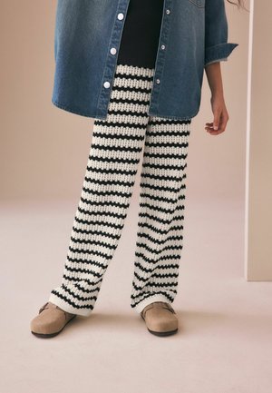 Person trägt schwarz-weiß gestreifte Strickhosen, beige Slipper, ein Jeanshemd und ein schwarzes Oberteil und steht drinnen.