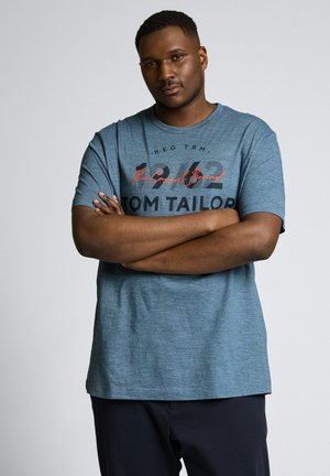 Mężczyzna ze skrzyżowanymi ramionami, ubrany w niebieski T-shirt z grafiką Tom Tailor i ciemne spodnie, stojący na jasnym, jednolitym tle.