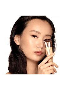 Eine Tube Foundation mit einem metallisch roségoldenen Deckel, die nahe dem Gesicht eines Models gehalten wird. Sie zeigt eine glatte, beige Textur und ein elegantes Design.