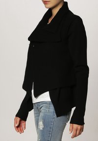 Svart, texturerad kardiagan med asymmetrisk fåll och bred krage. Har långa ärmar och en subtil accent nära kragen.