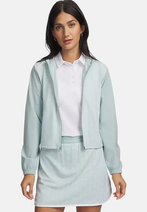 Vrouw draagt een lichtblauwe jas met ritssluiting over een witte blouse met kraag en bijpassende lichtblauwe rok op een effen achtergrond.