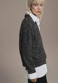Maglione oversize grigio scuro con lavorazione a maglia, indossato sopra una camicia bianca con colletto. Pantaloni neri e uno sfondo neutro completano il look.