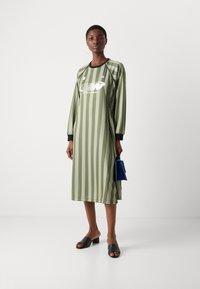 Olivenfarbenes Midikleid mit vertikalen Streifen, langen Ärmeln, Rundhalsausschnitt und sichtbarem Logo. Kombiniert mit schwarzen Blockabsatz-Sandalen.