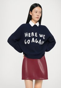 Marineblå striktrøje med hvid tekst "HER KØRER VI IGEN" parret med en glansfuld burgunder faux læder miniskirt, lagd over en skjorte med krave.