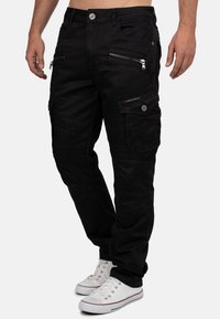 Pantaloni cargo neri in denim, dotati di multiple tasche con zip, design aderente e cuciture a vista. Abbinati a sneakers bianche.