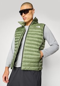 Groene puffer bodywarmer met een ritssluiting aan de voorkant, horizontale stiksels, hoge kraag en merklogo. Draag een grijze langsleevenshirt eronder.