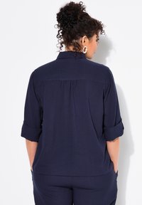 Camisa de color azul marino, de mangas cortas, con cuello tipo polo y puños enrollados, que presenta un pliegue en la parte posterior y un ajuste relajado. Tejido ligero.