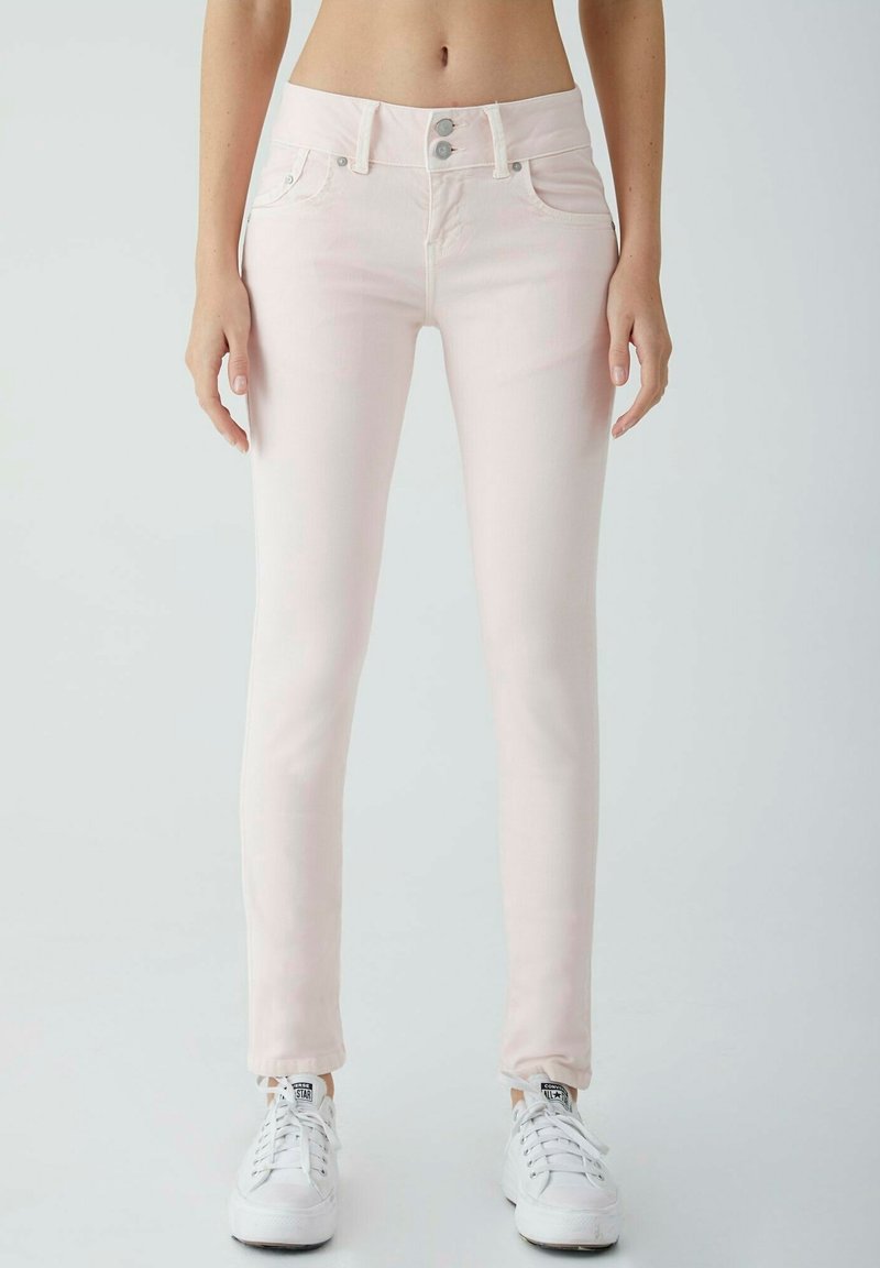 Jean slim rose clair avec fermeture double bouton, porté avec des baskets Converse blanches par une personne debout devant un fond uni.