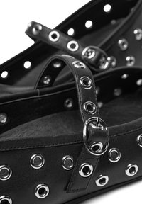 Zapatos planos negros de cuero con ojales plateados y correas. Presentan una punta redondeada y una textura suave.