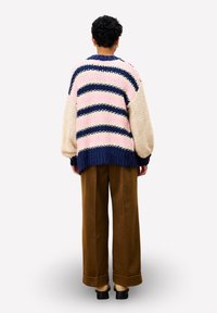 Person mit kurzen lockigen Haaren, die einen cremefarbenen, rosa und marineblauen gestreiften, grob gestrickten Pullover, braune weite Hosen und schwarze Schuhe trägt und mit dem Rücken steht.