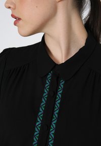 Zwarte blouse met een kraag, knoopsluiting en kleurrijke geometrische borduursels in groen en blauw aan de voorkant.