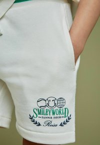 Witte katoenen shorts met een afbeelding van een glimlachend gezicht, tennisballen en een wereldbol. De tekst luidt "SMILEYWORLD Tennis Tour" in groen en marineblauw.