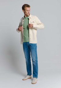 Beige button-up shirt over een groene T-shirt, gecombineerd met blauwe jeans en lichte sneakers. Het shirt heeft twee voorzakken.