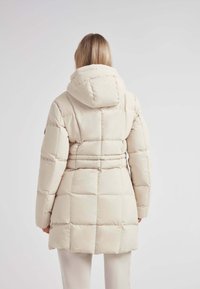 Abrigo de plumas beige con capucha, que presenta un diseño acolchado, cintura ajustada y mangas largas. Textura suave y estética minimalista.