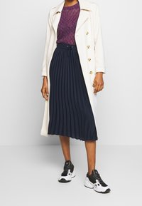Un trench coat bianco sopra un top viola testurizzato, abbinato a una gonna plissettata blu navy e sneakers nere con dettagli bianchi.