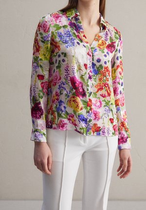 Frau trägt eine langärmelige Bluse mit mehrfarbigem Blumenmuster und weißen Hosen vor einem schlichten Hintergrund.