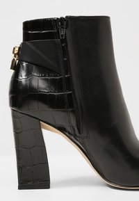 Bottine en cuir noir avec texture crocodile sur le talon et le dos, fermeture éclair latérale et détail de boucle dorée.