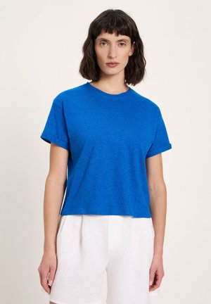 Frau mit kurzen dunklen Haaren, die ein blaues, kurzärmeliges Hemd und weiße Shorts mit hoher Taille trägt, steht vor einem schlichten hellen Hintergrund.