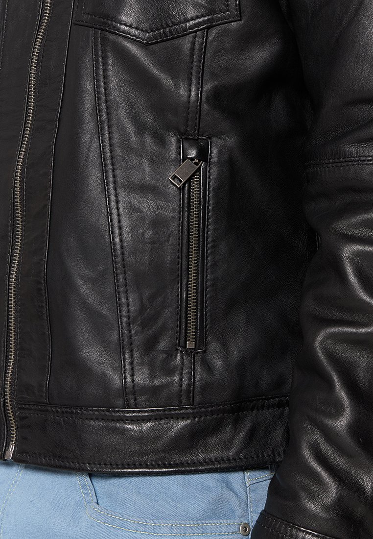 Primo piano di una giacca in pelle nera con una tasca verticale con zip e dettagli di cucitura, indossata su jeans di denim azzurri.