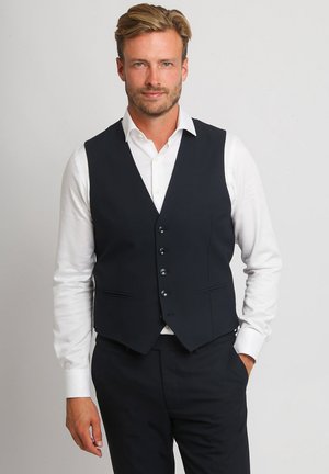 Man draagt wit overhemd en zwarte op maat gemaakte vest en broek, staat met één hand in zijn zak tegen een effen achtergrond.