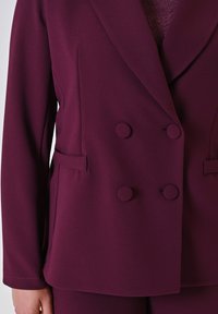 Frau trägt ein doppelt geknöpftes Blazer in Pflaumenfarbe mit mit Stoff überzogenen Knöpfen und passenden Hosen, die Hand entspannt an ihrer Seite.