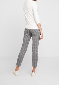 Pantaloni grigi a scacchi con una vestibilità slim, abbinati a un top bianco a maniche lunghe. Il modello è in piedi su uno sfondo bianco, indossando sneakers bianche.